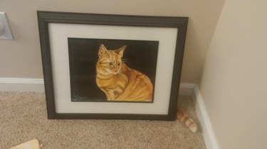 Cat sitting behind a painting of itself with just its tail visible