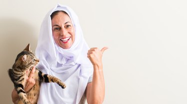 Arabian woman wearing white hijab holding a cat pointing with hand and finger up with happy face smiling