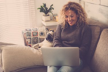 beautiful blonde curly woman working at home with a laptop internet connected while her best friends creamy pug look at the screen to check the work - friendship and lovely puppy concept
