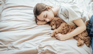 Young woman is lying and sleeping with poodle dog in bed.