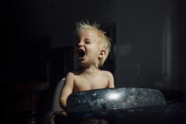 Shirtless baby boy screaming while enjoying breeze from air conditioner in darkroom at home