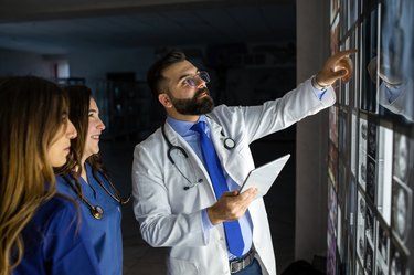 A doctor wearing a lab coat and stethoscope examines X-rays as two doctors wearing scrubs stand nearby.