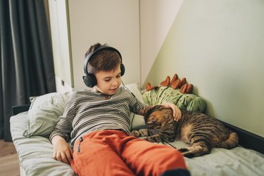 A happy child in a white T-shirt lies on a white bed with a phone and headphones in his ears. The boy listens to music on the phone. Modern children. Children and modern gadgets. A child's smartphone.