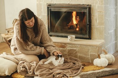 Hand caressing cute cat on cozy blanket at fireplace close up, autumn hygge. Stylish female in warm sweater together with kitty relaxing at fireplace in rustic farmhouse.