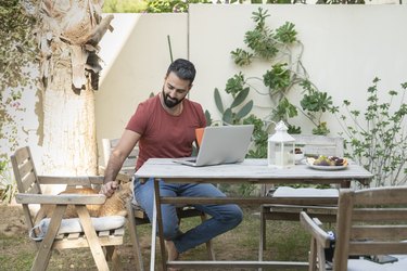 Smiling man with laptop stroking cat sitting on chair in garden