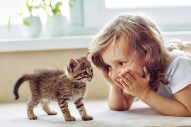 A little girl is laying on her stomach with her hands on her face. In front of her is a small kitten.