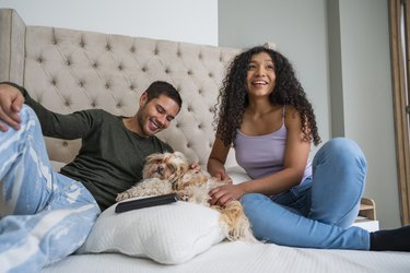 A Latino couple are lying on their bed with their pet long-haired small dog and with their tablet watching a movie while spending time together