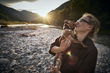 Woman cuddling with dog at a riverbank at sunset