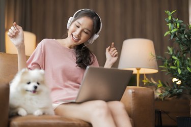 young asian cheerful freshness Woman waving while she is conversation during video call and wear headphone device using  laptop while her white hair colour pomeranian dog pet is relax laying down sit next to her on sofa armchair at home