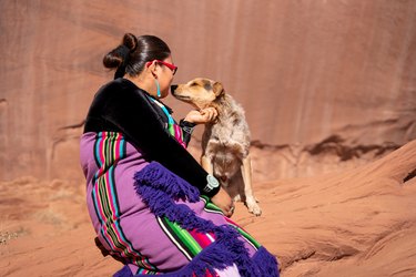 Young Navajo Native American Woman in her early twenties in traditional clothing sitting Playing with her Dog in Monument Valley