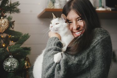 Merry Christmas! Woman in cozy sweater hugging cute cat near stylish christmas tree with vintage baubles. Pet and winter holidays. Adorable kitty and happy woman cuddling in festive room