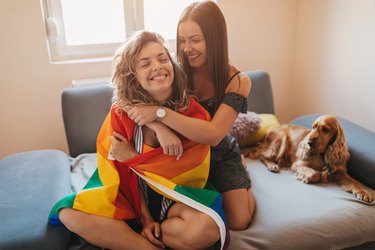 woman wrapped in rainbow flag with other woman hugging her. both women are smiling. a cute spaniel dog is on the couch behind them.