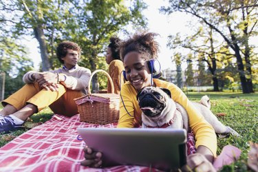 Beautiful young family lying on a picnic blanket with their dog, enjoying an autumn day in park while using digital tablet.