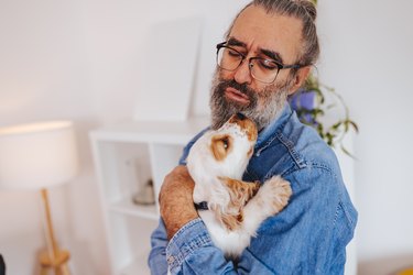 a man with a beard and glasses tenderly holds a small white puppy who licks his face