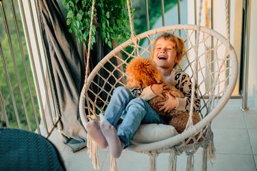 happy little girl, child hugging with a smile her pet, poodle dog at home on the balcony in spring, summer in a cotton-fringed hammock chair at sunset.