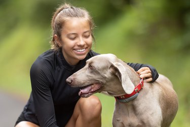 Smiling teen girl pets her Weimaraner during a walk in the nature.