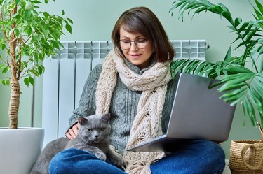 Middle-aged woman in warm clothes with cat using laptop at home
