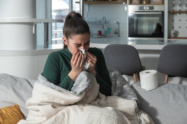Young ill woman wrapped in blanker blowing her nose in to the toilet paper. Sick female sitting at home on sofa try to warm up suffering from common cold or flu.