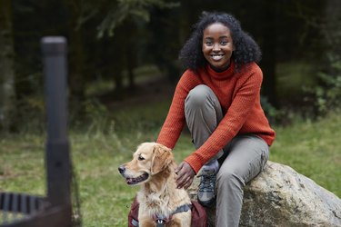 Woman sitting on rock with dog in forest, wearing red sweater and smiling at the camera