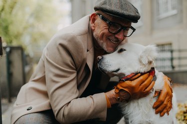 Happy senior man sitting on bench and embracing his dog outdoors in park in city.