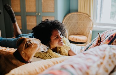 Beautiful woman lies on her stomach on a colourful bed, beside her lurcher dog, and takes a selfie.