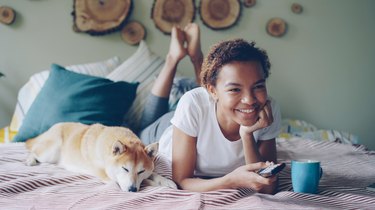 African American woman is watching TV holding remote control pressing buttons and laughing while her pet dog is lying beside her. Entertainment and technology concept.