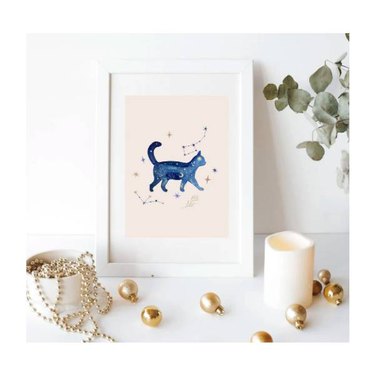 A digital file of a cat surrounded by constellations in watercolor style that's been printed out and framed