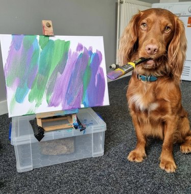 dog standing next to a painting with a paintbrush in their mouth.