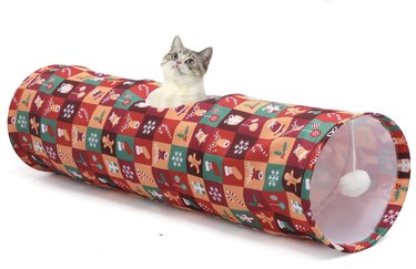 Cat playing in a Christmas-patterned pop-up tunnel with a pompom on a string at one end and a peep hole.