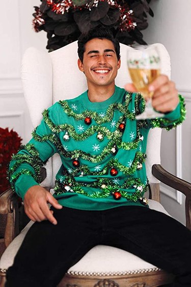 Man wearing green sweater with green tinsel, red and gold ornaments, and white snowflakes.