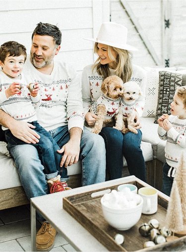 Two parents, two children, and two dogs wearing matching cream-colored holiday sweaters with black and red motifs.