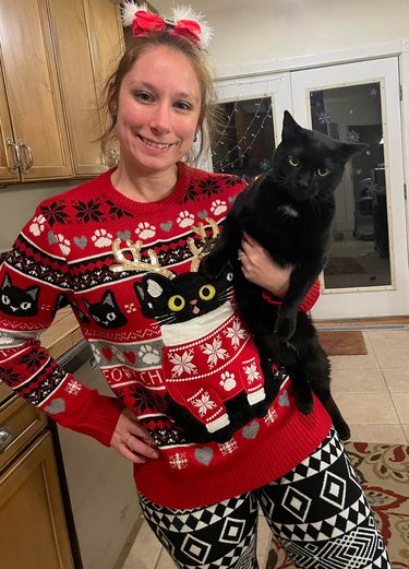 A woman is wearing a Christmas sweater with a black cat on it, while holding her own black cat.