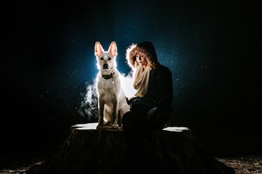 Woman in a hooded jacket sits beside a white shepherd dog against a dark background.