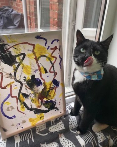 A small painted canvas resting in a windowsill. Next to the painting sits a cat with their tongue out, licking their nose.
