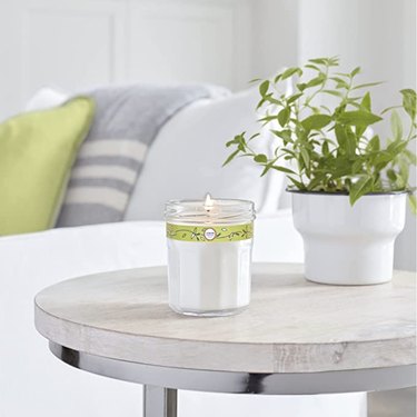 a white candle in a clear glass container on a wood side table with a potted plant next to a white couch