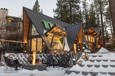 Modern A-frame with numerous peaks and a black metal roof. Below is a sunken seating area with a fire pit and people sitting around it.