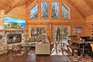 Log cabin living room with vaulted ceilings, a wall of windows, a stone fireplace, large TV, sectional, and dining table.