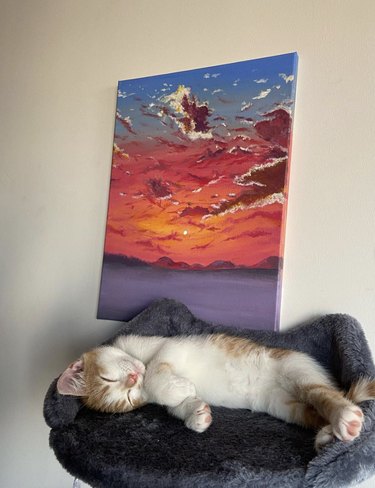 An orange and white kitten sleeping in an elevated cat bed with a canvas painting of a sunset behind them on a wall.