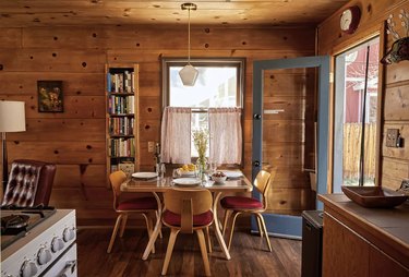 Knotty pine cabin interior showing a dining table, a bookcase, and an open glass door leading to the backyard.