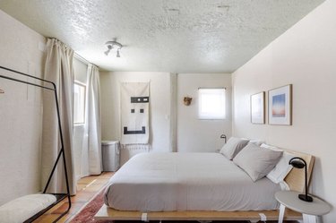 The bedroom area of the studio featuring a platform bed with gray bedding, a freestanding clothing rack, two windows, and wall art.