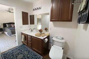The bathroom of the private room with a walnut-colored vanity with a single sink, a toilet, and storage. The bathroom door is open, providing a glimpse of the carpeted bedroom with a ceiling fan.