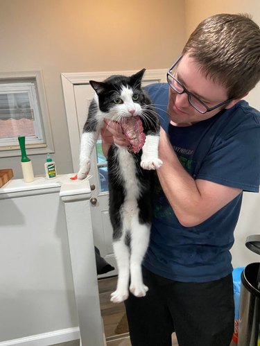 Man holding cat with piece of meat hanging from its mouth