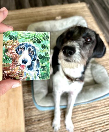 Person holds mini canvas portrait, and dog is in background.