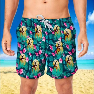 A man on a beach wearing floral print and blue swim shorts with a Golden Retriever's face printed on it.