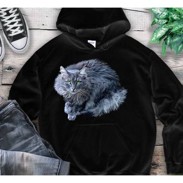 A black hooded sweatshirt with a realistic illustration of a fluffy grey cat on it.