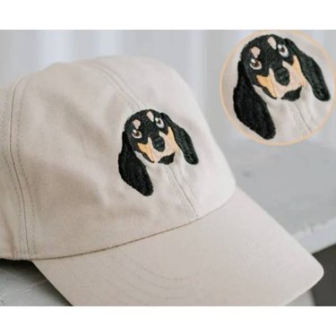 A cream hat with a brown and white dog's face embroidered on the front of it, alone with a close up of the embroidered image