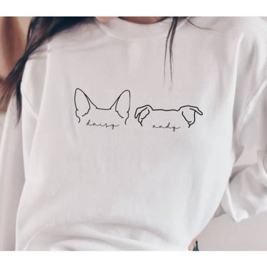 A woman wearing a white crewneck sweatshirt with the outlines of dogs' ears on it and their names personalized under the images.