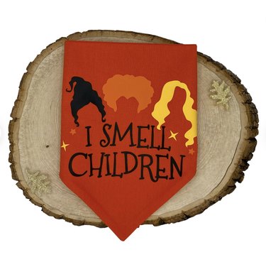 Reddish orange dog bandanna with the hair silhouettes of the Sanderson sisters and the words "I smell children."