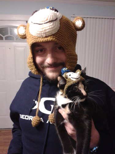 Man wearing crocheted monkey hat holds cat with matching hat
