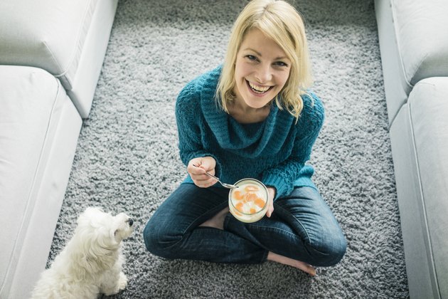 Portrait of happy woman with dog eating fruit yoghurt in living room